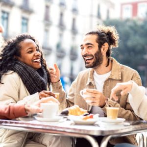 Four friends laugh together while enjoying coffee and pastries at an outdoor café table.