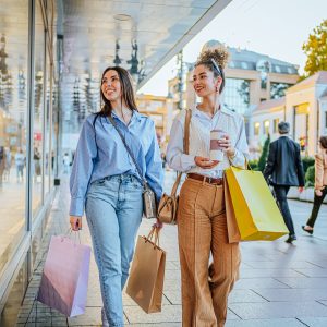 Two smiling women carry shopping bags and coffee while strolling past storefronts on a city sidewalk